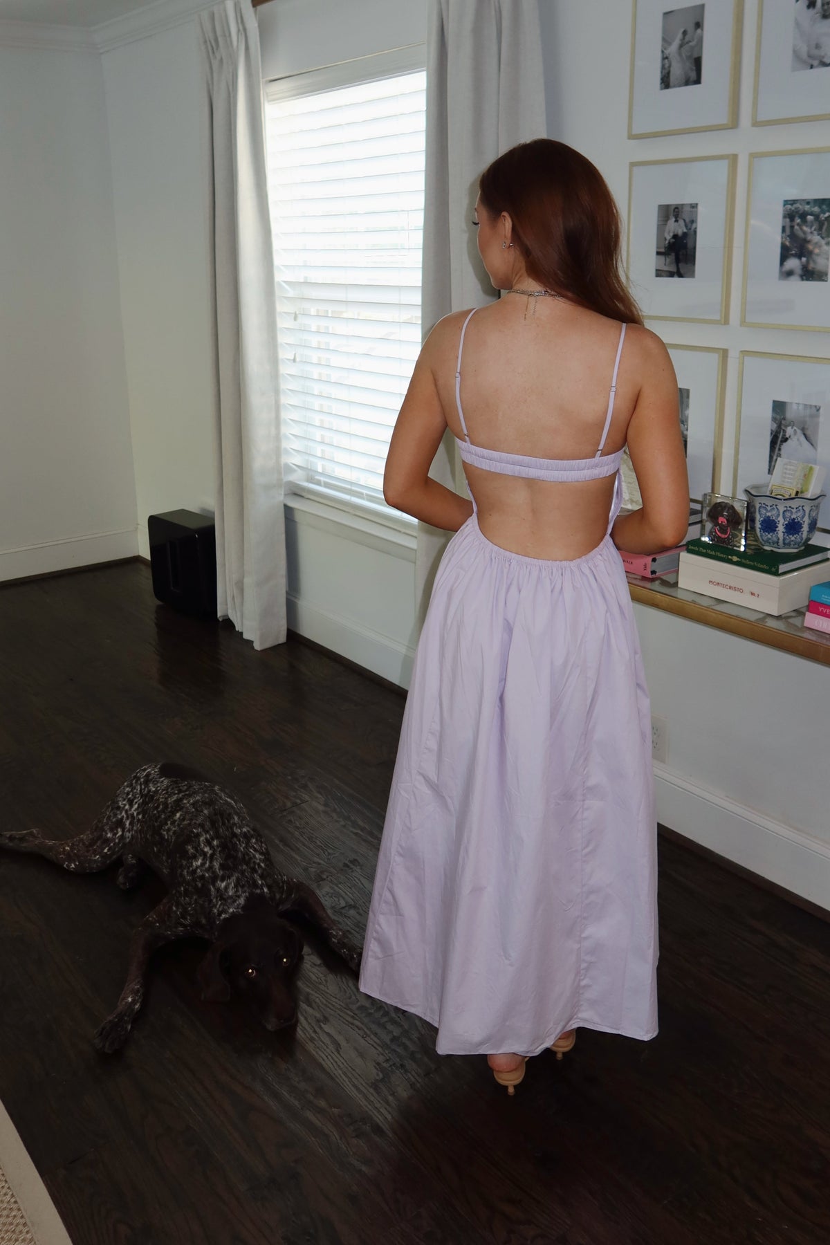 A woman stands with her back to the camera in a bright room, wearing a light lavender backless maxi dress with spaghetti straps. A dark dog is lying on the dark wood floor nearby, and framed photos and a shelf are visible on the wall behind her.
