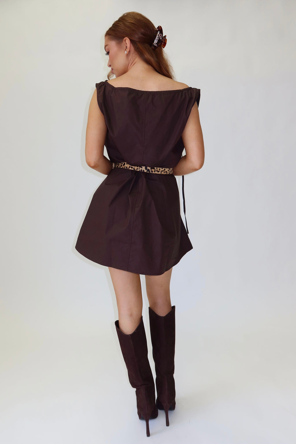 A woman is shown from behind wearing a dark brown off-the-shoulder mini dress with a gold patterned belt and matching dark brown knee-high boots, standing against a white studio background.