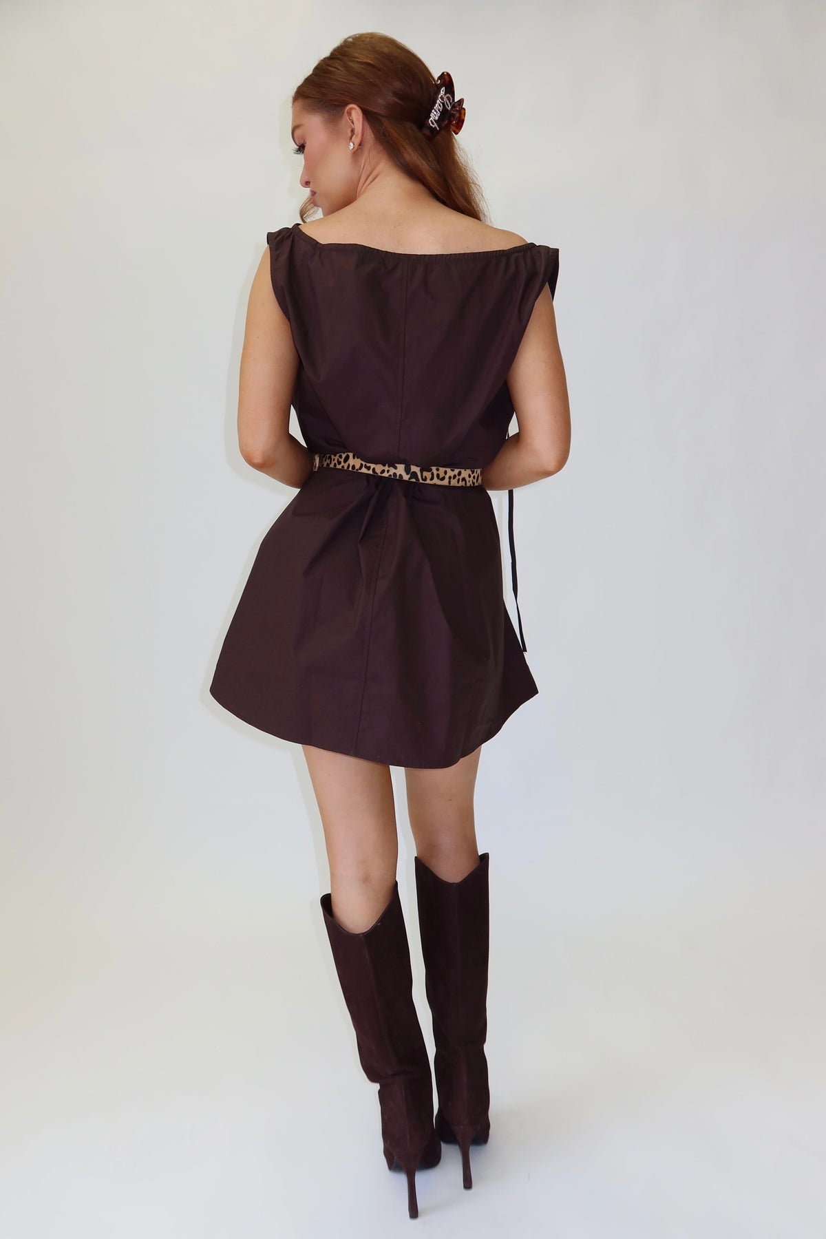 A woman is shown from behind wearing a dark brown off-the-shoulder mini dress with a gold patterned belt and matching dark brown knee-high boots, standing against a white studio background.