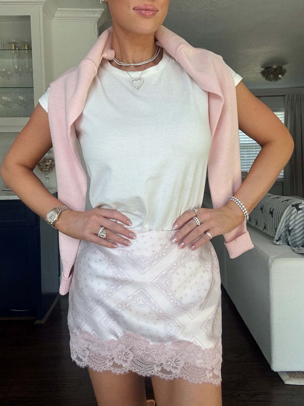 A woman posing indoors wearing a white crewneck t-shirt, a light pink blazer draped off her shoulders, and a white lace mini skirt with a pink lace hem.