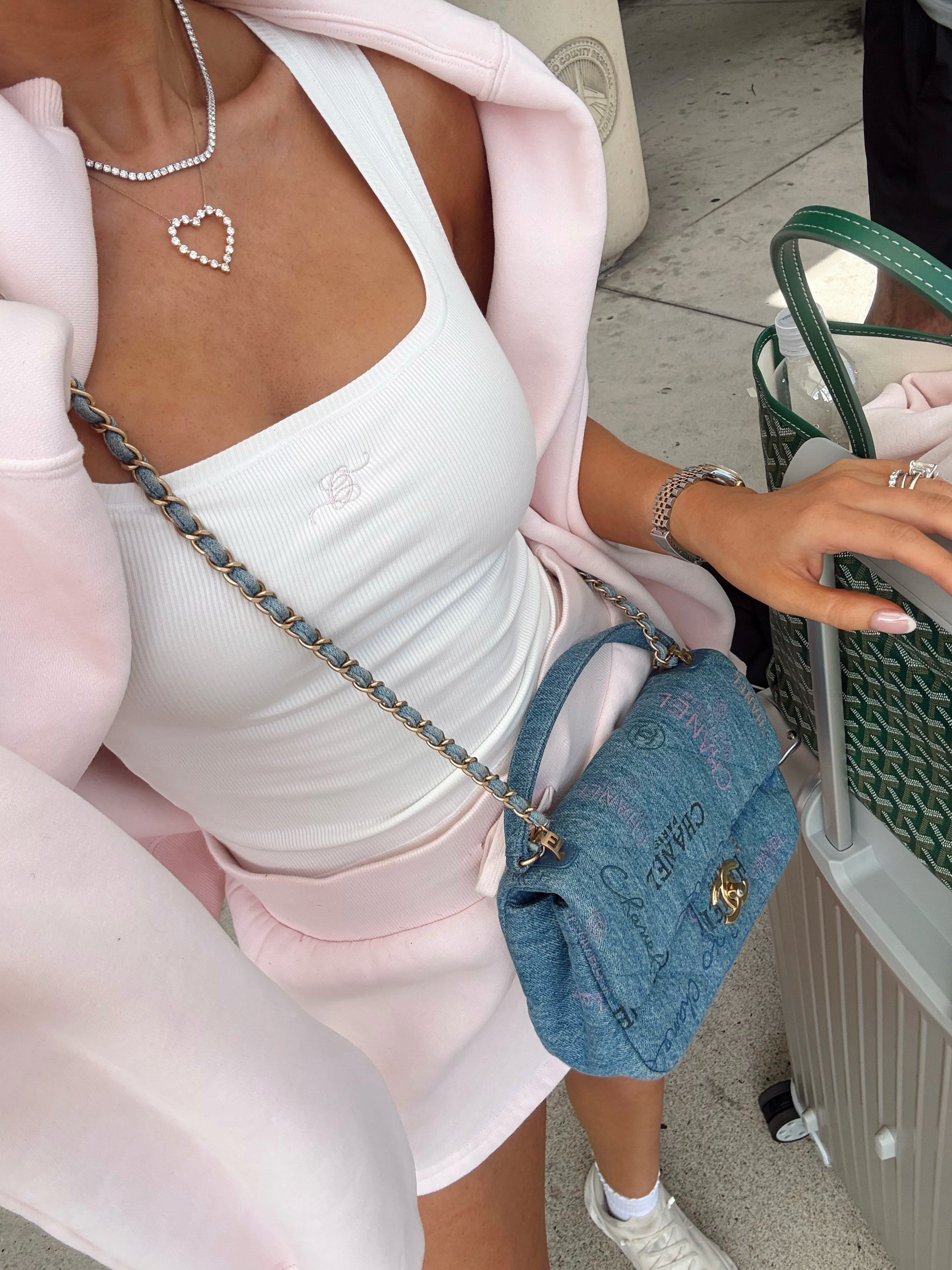 A person wearing a white ribbed tank top and light pink hoodie, carrying a blue textured shoulder bag with a chain strap. The person is also wearing a heart-shaped rhinestone necklace and a bracelet, standing next to a green patterned tote bag and a suitcase.