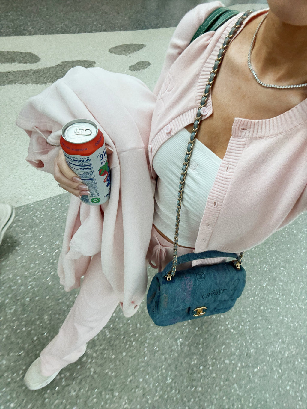 A person wearing a light pink cardigan and matching pink pants, holding a pink garment in one hand while carrying a blue Chanel crossbody bag and a can of soda. The background is a gray speckled floor.