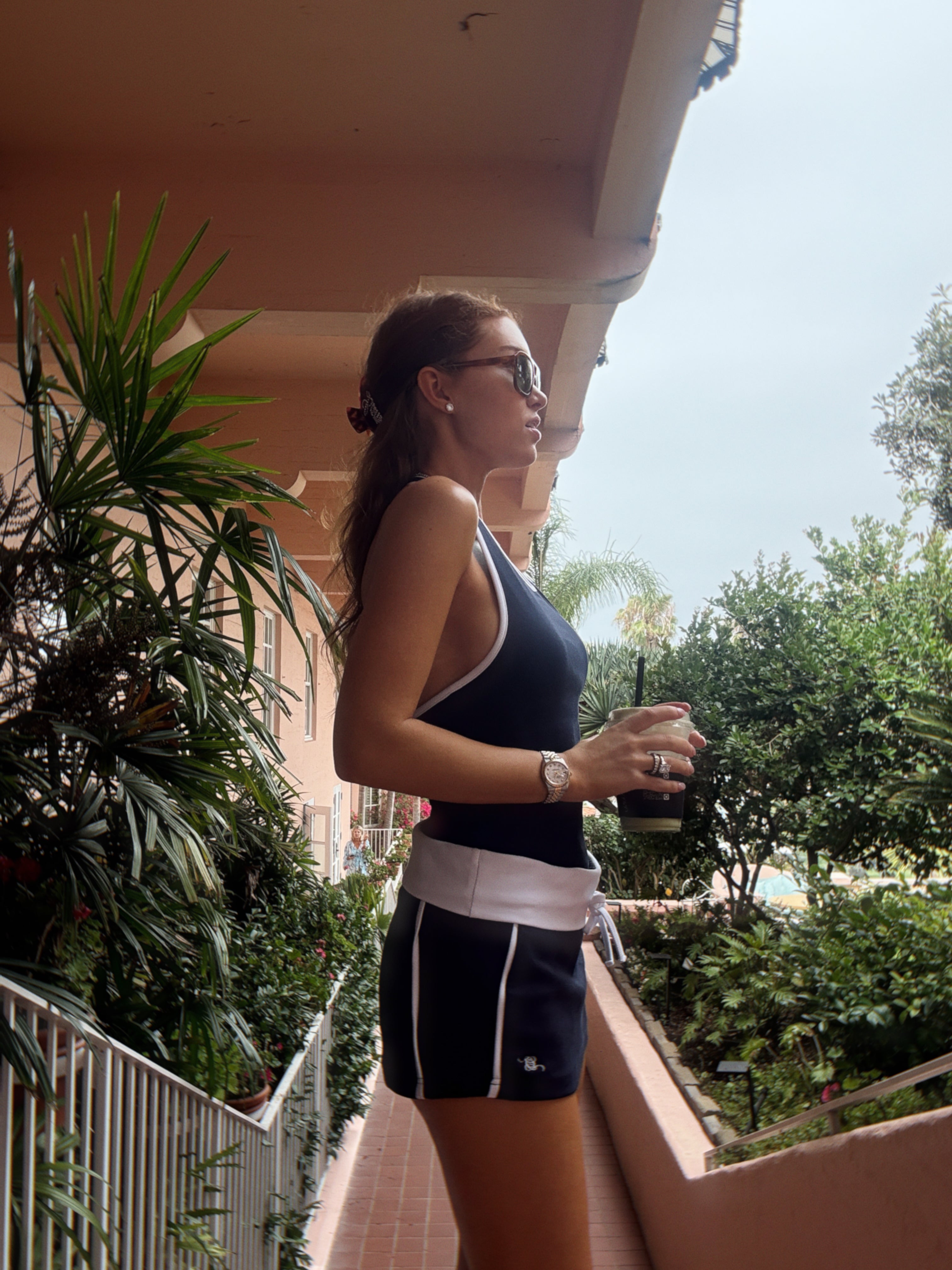 A woman wearing a navy blue tank top and matching navy shorts with white trim stands on an outdoor balcony with green plants and a terracotta wall.
