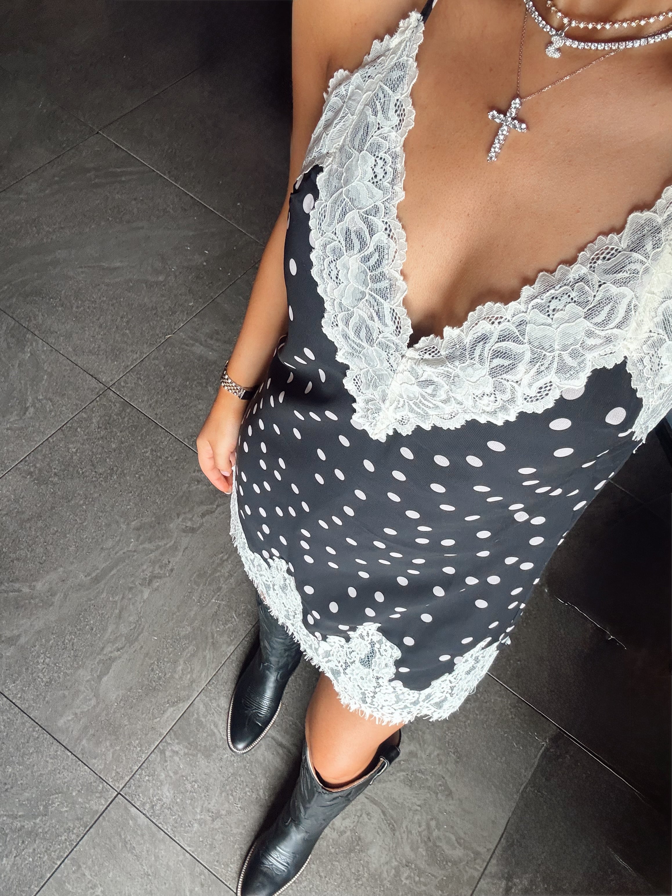 A woman wearing a black satin slip dress with white polka dots and a white lace V-neck trim, paired with black pointed-toe ankle boots and layered silver necklaces, standing on a dark gray tiled floor.