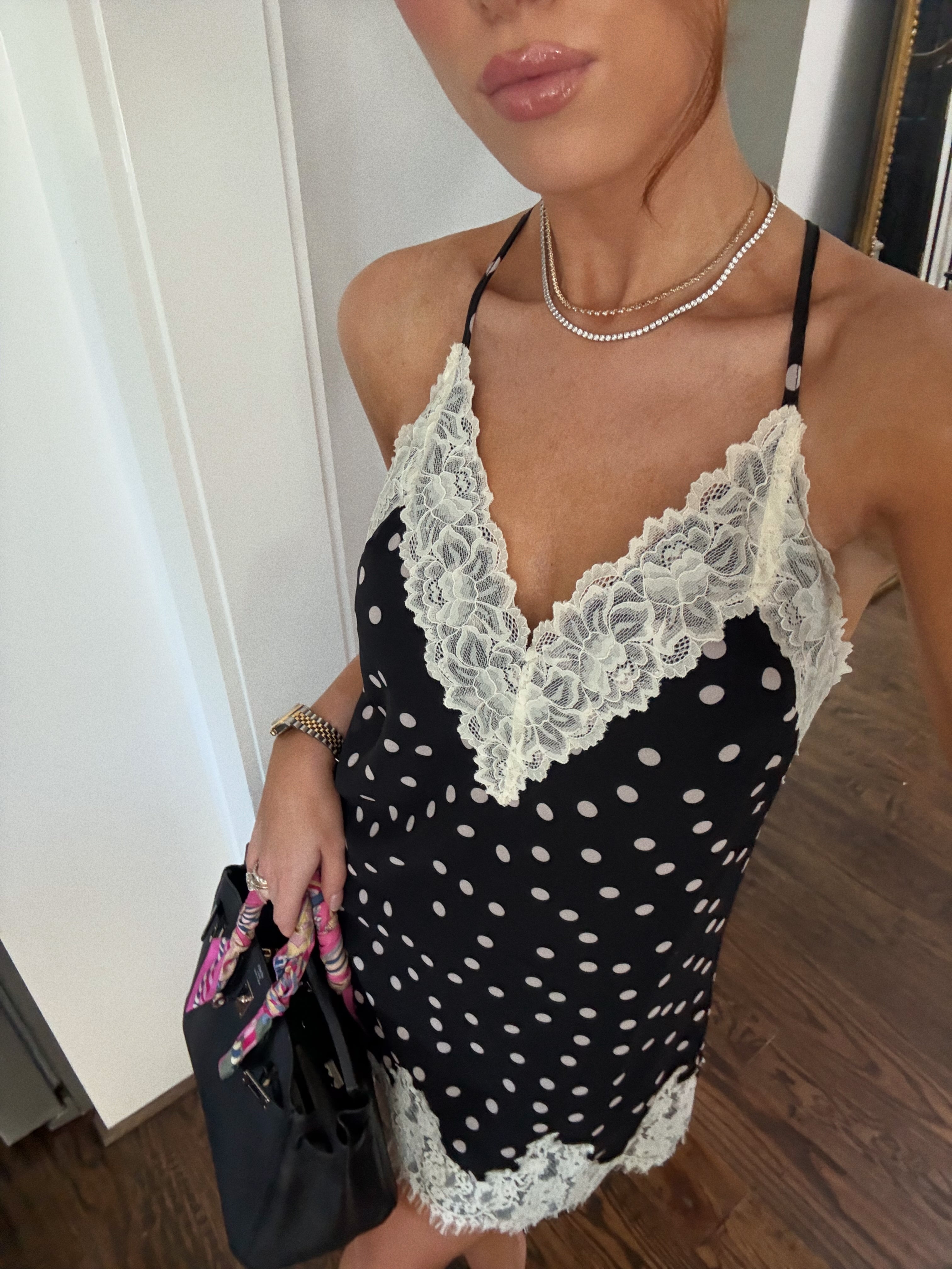A woman wearing a black camisole-style dress with white polka dots and white lace trim, photographed indoors against a light wall and wood floor.