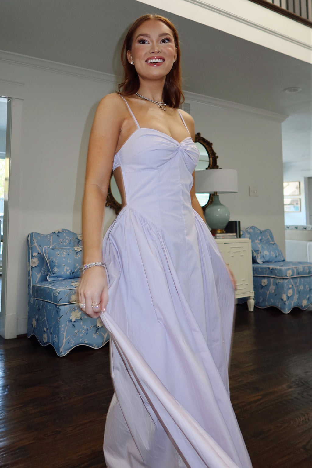 A woman wearing a lavender spaghetti-strap formal maxi dress with a sweetheart neckline, photographed in a spacious home interior with hardwood floors and blue upholstered chairs.
