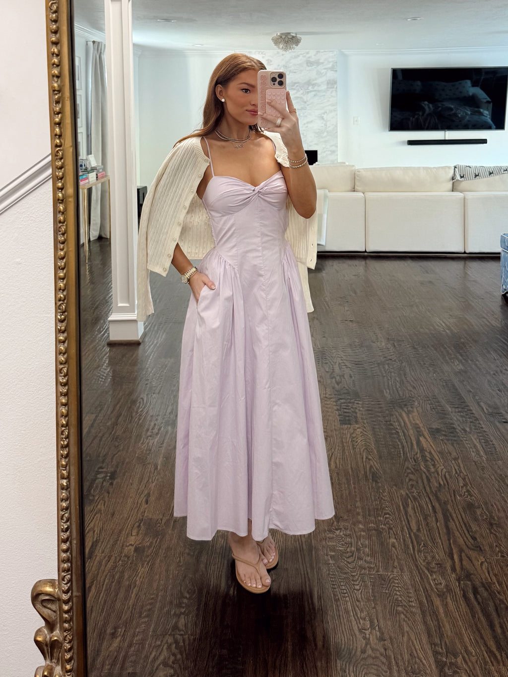 A woman takes a mirror selfie in a home wearing a light lavender formal dress with a sheer white cape. The setting is a modern home with dark wood floors, a white living room, and a large ornate gold-framed mirror.