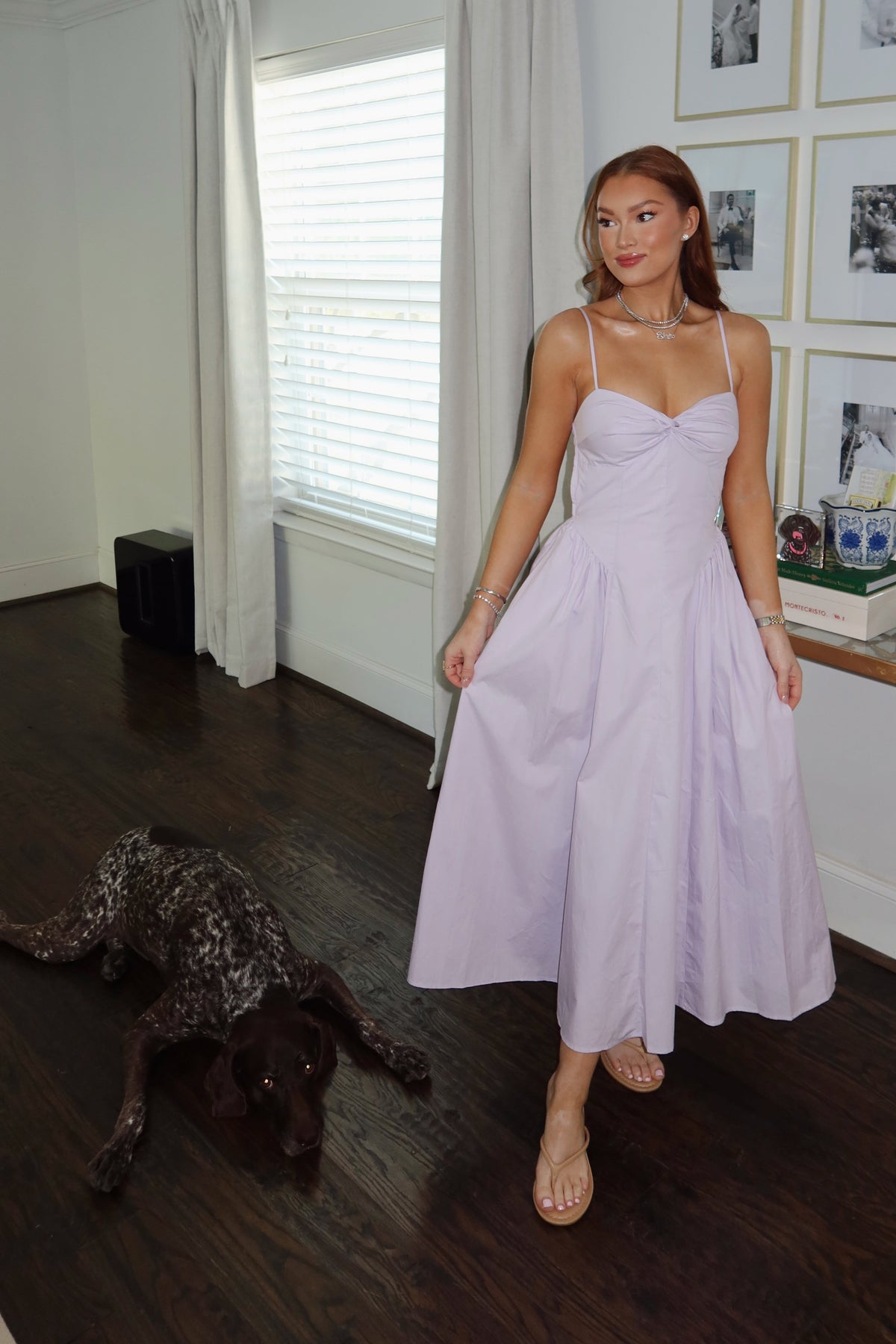 A woman wearing a light lavender midi dress with spaghetti straps and a sweetheart neckline stands in a room with dark wood floors, white walls, and framed pictures. A dark-colored german shorthaired pointer dog is lying on the floor to the left.