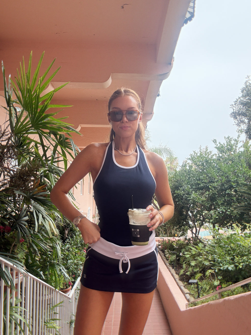 A woman wearing a navy blue athletic tank top and matching shorts with a white waist detail, holding a coffee cup in an outdoor setting with plants and a pink building in the background.