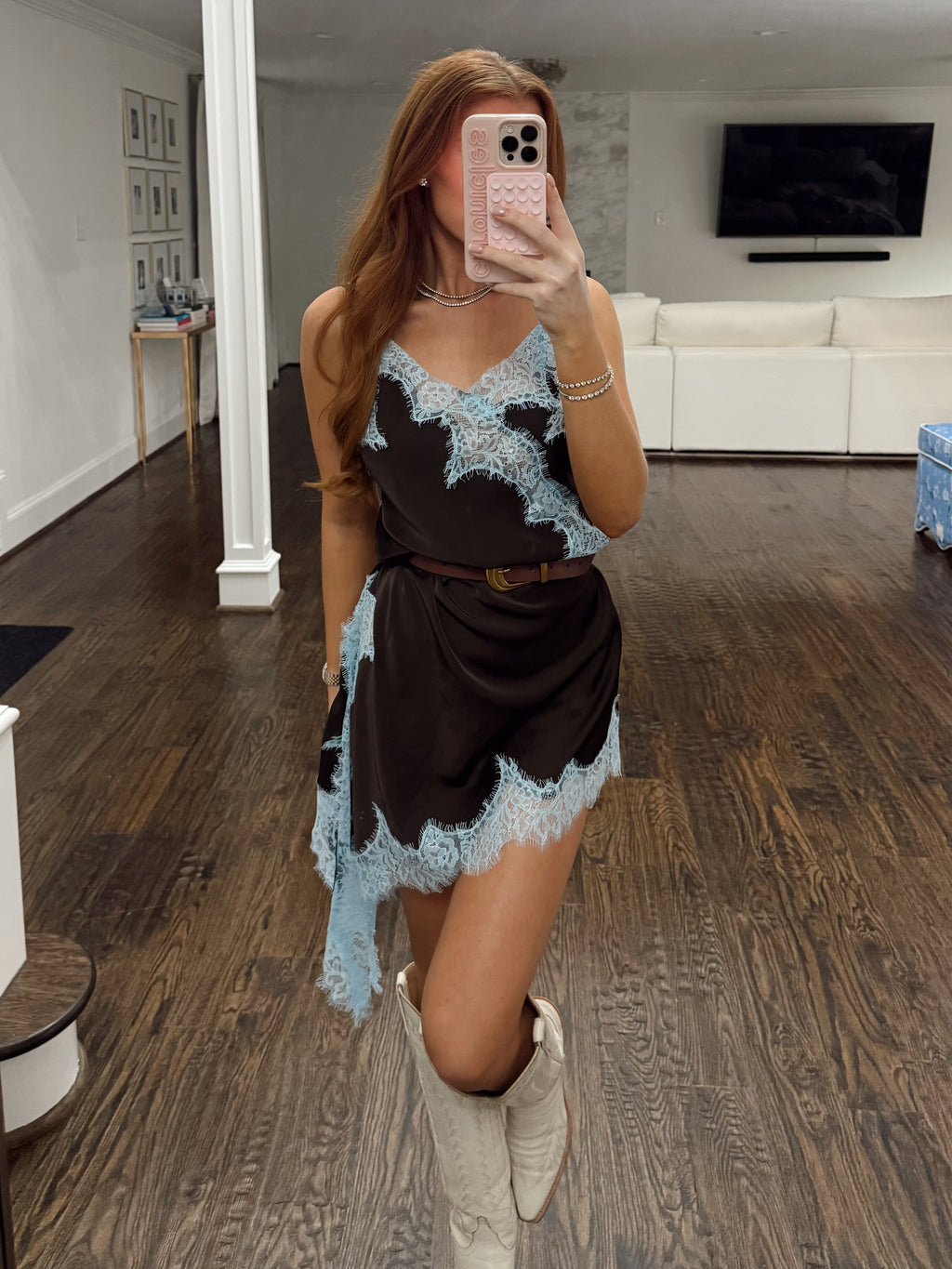 A woman takes a mirror selfie in a modern room with dark wood floors, wearing a brown satin-like mini dress with light blue lace trim and white cowboy boots.