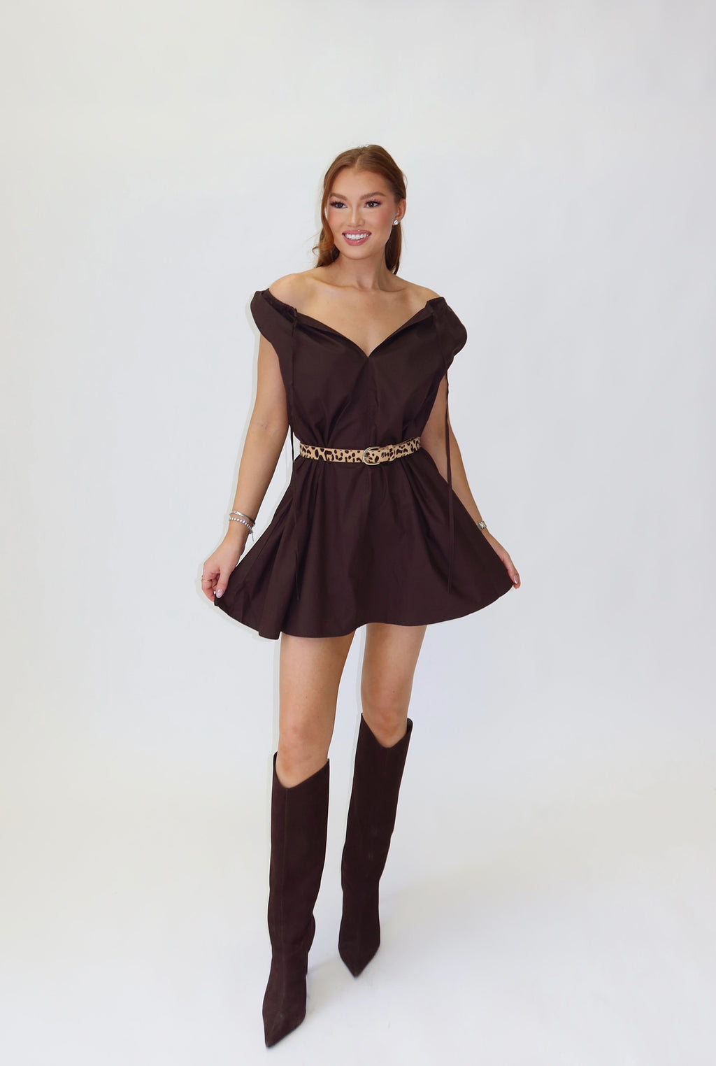 A woman modeling a dark brown off-the-shoulder mini dress with a gold metallic belt, paired with matching dark brown knee-high boots, standing against a white studio backdrop.