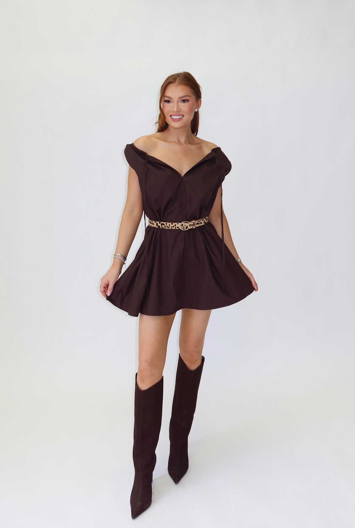 A woman modeling a dark brown off-the-shoulder mini dress with a gold metallic belt, paired with matching dark brown knee-high boots, standing against a white studio backdrop.