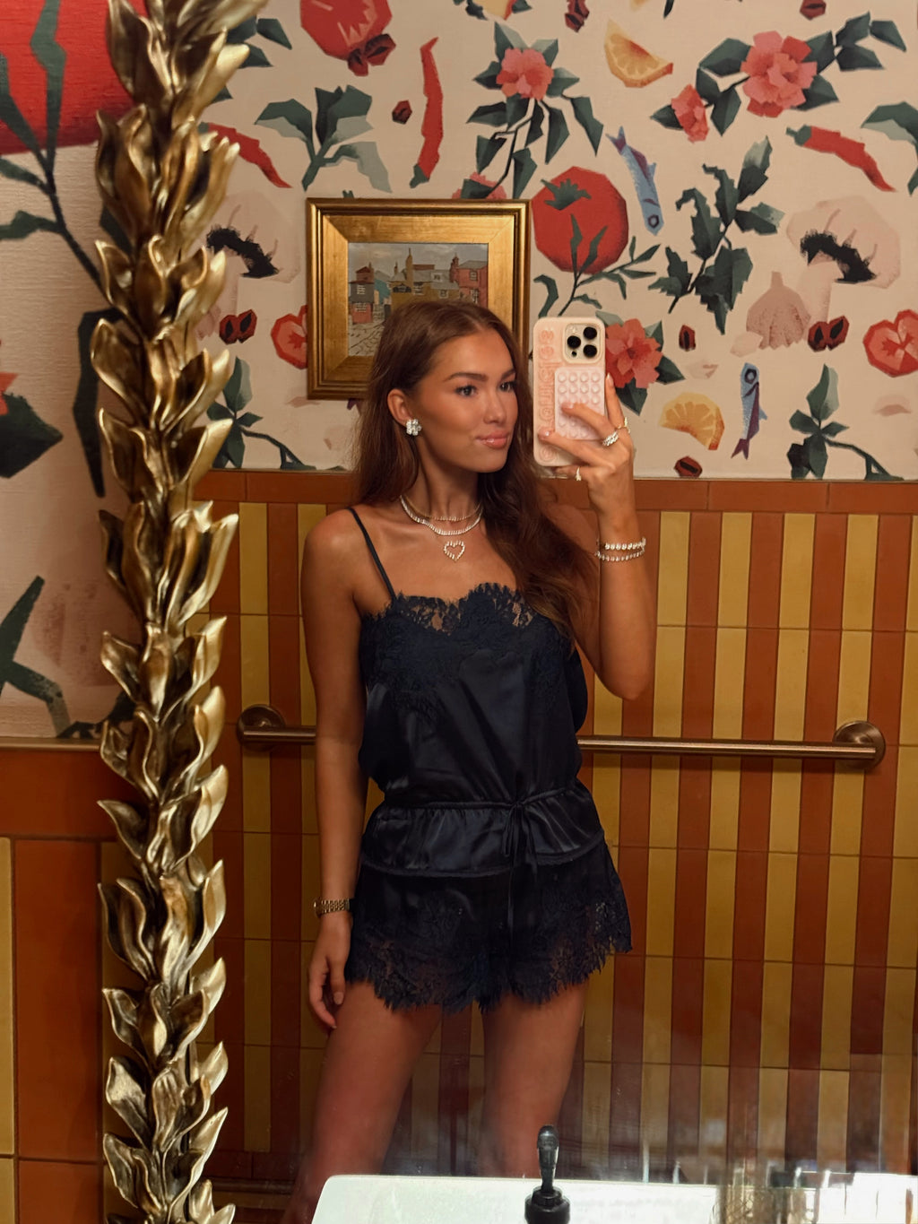 A woman takes a mirror selfie wearing a black satin camisole top with lace trim and a matching black lace mini skirt. The setting features a gold decorative mirror frame on the left, striped wood paneling, and a floral wallpaper background.