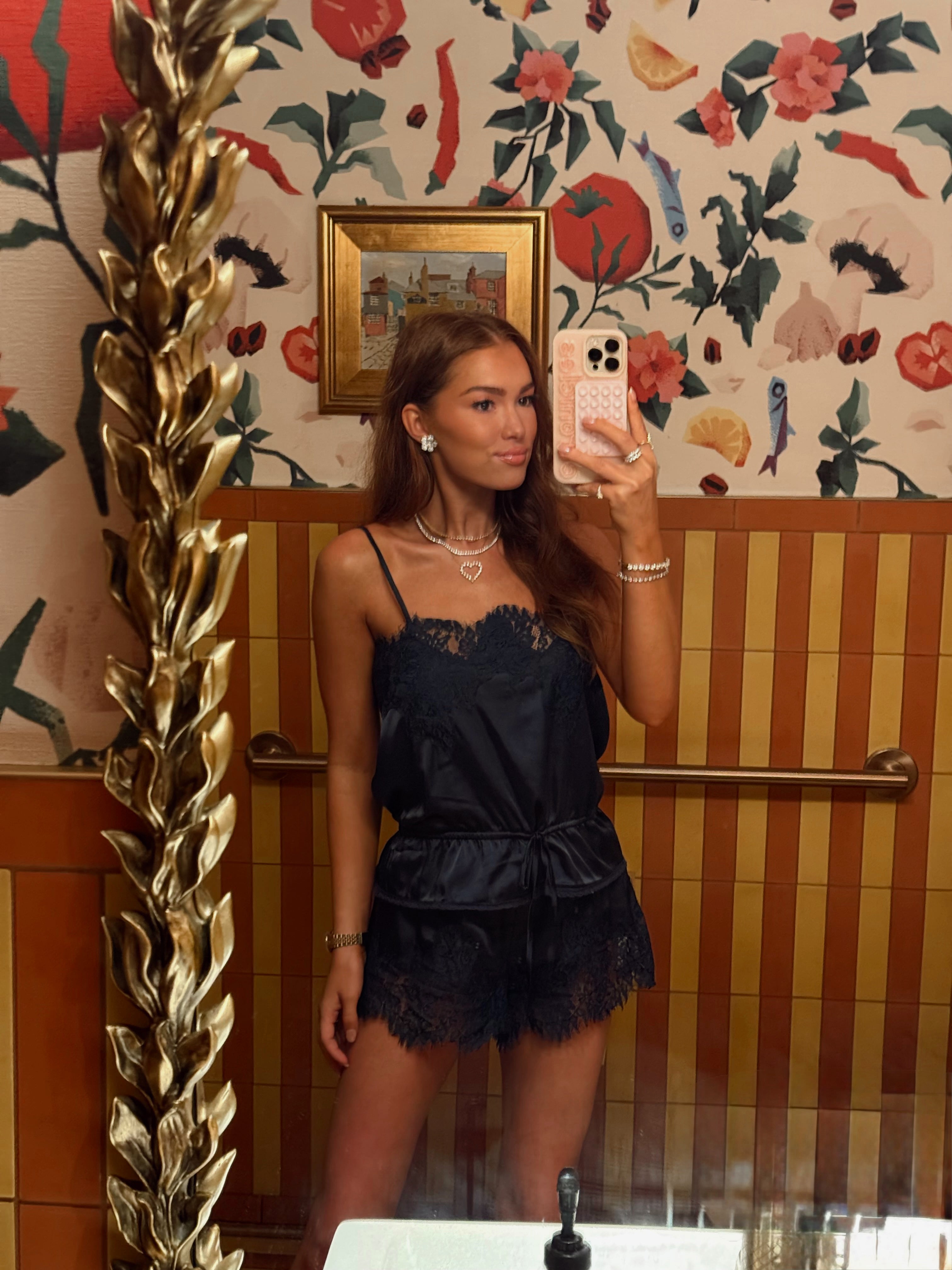 A woman takes a mirror selfie wearing a black satin camisole top with lace trim and a matching black lace mini skirt. The setting features a gold decorative mirror frame on the left, striped wood paneling, and a floral wallpaper background.