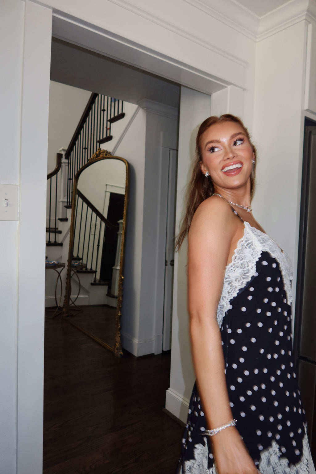 A woman wearing a navy blue and white polka dot dress with white lace trim, standing in a doorway of a home interior with a staircase in the background.