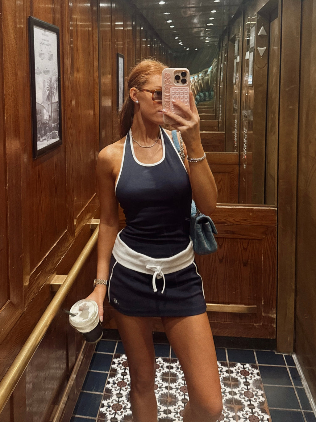 A woman takes a mirror selfie in a wood-paneled hallway, wearing a navy blue athletic tank top and matching shorts with white trim and a white drawstring waist.