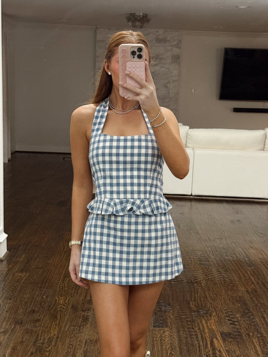 A woman takes a mirror selfie in a living room wearing a blue and white gingham halter mini dress with a ruffled waist. The background shows a white sectional sofa, a dark wood floor, and a wall-mounted television.