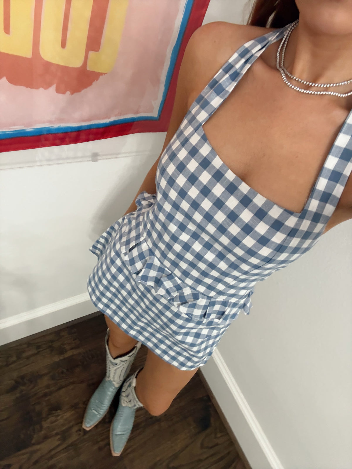 A person wearing a blue and white gingham halter mini dress with a ruffled hem, paired with light blue western boots and a silver beaded necklace, standing in a room corner.