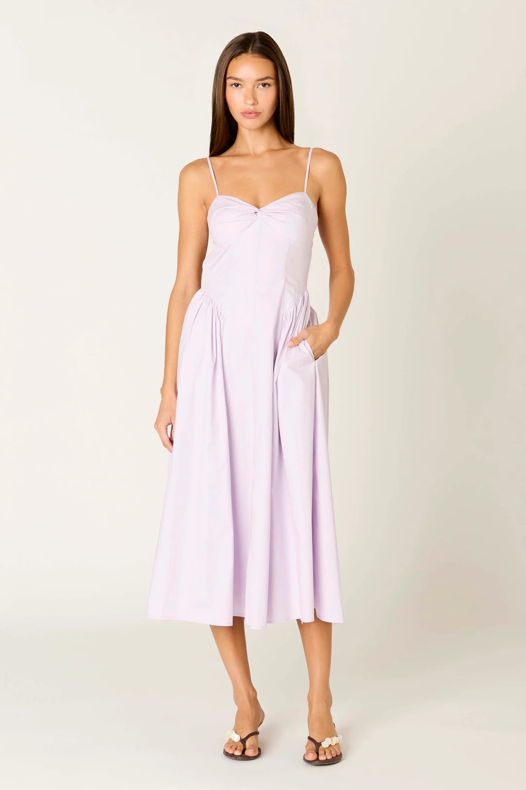 A woman modeling a light lavender midi dress with spaghetti straps, a sweetheart neckline, and side pockets, set against a plain light beige studio background.
