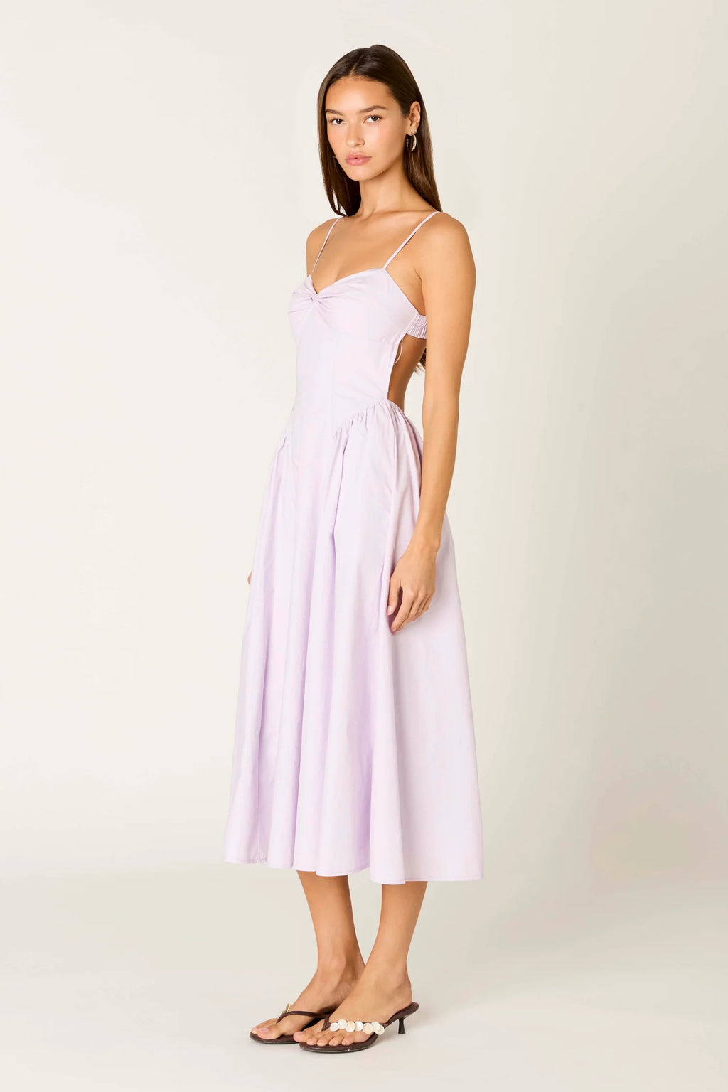A woman modeling a light lavender midi dress with thin spaghetti straps, a sweetheart neckline, and side cutouts, paired with black heeled sandals, against a plain off-white studio background.
