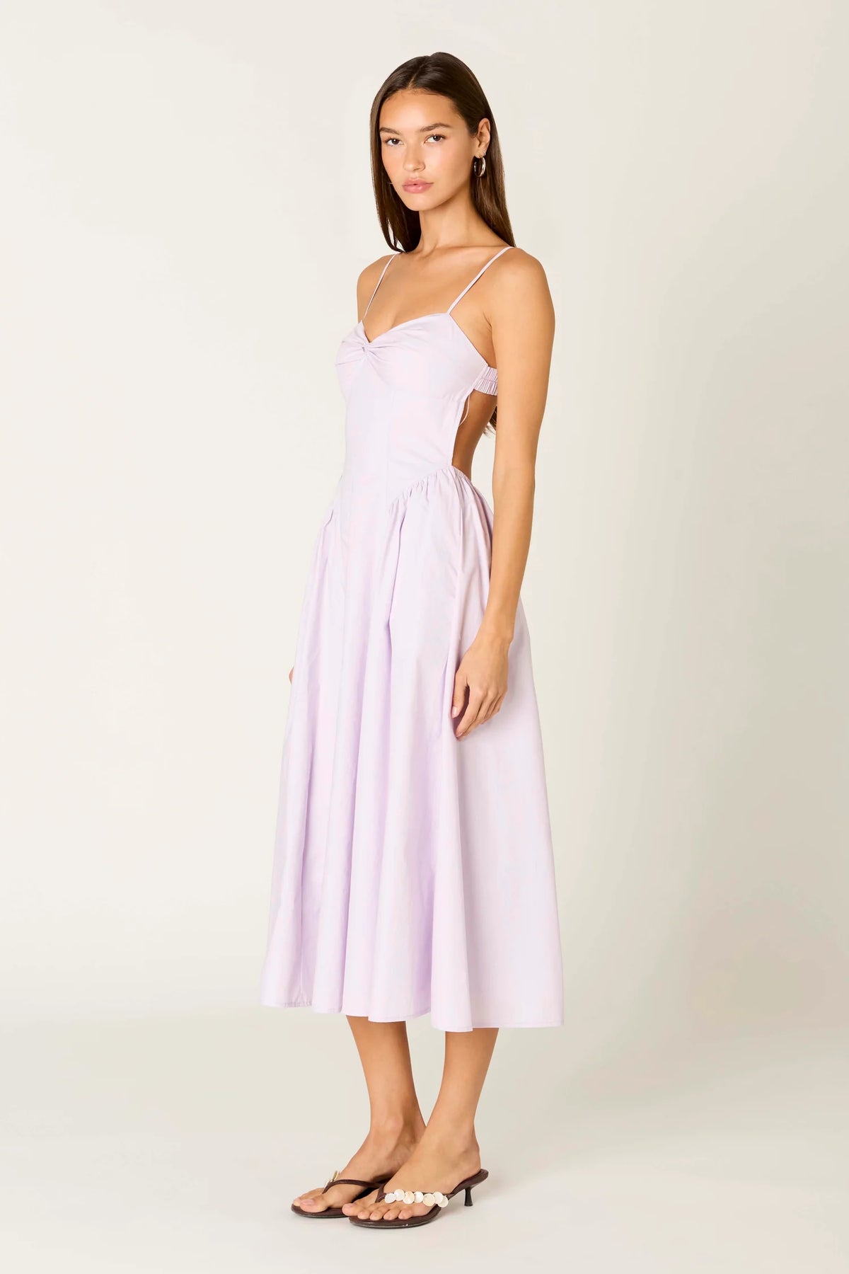A woman modeling a light lavender midi dress with thin spaghetti straps, a sweetheart neckline, and side cutouts, paired with black heeled sandals, against a plain off-white studio background.