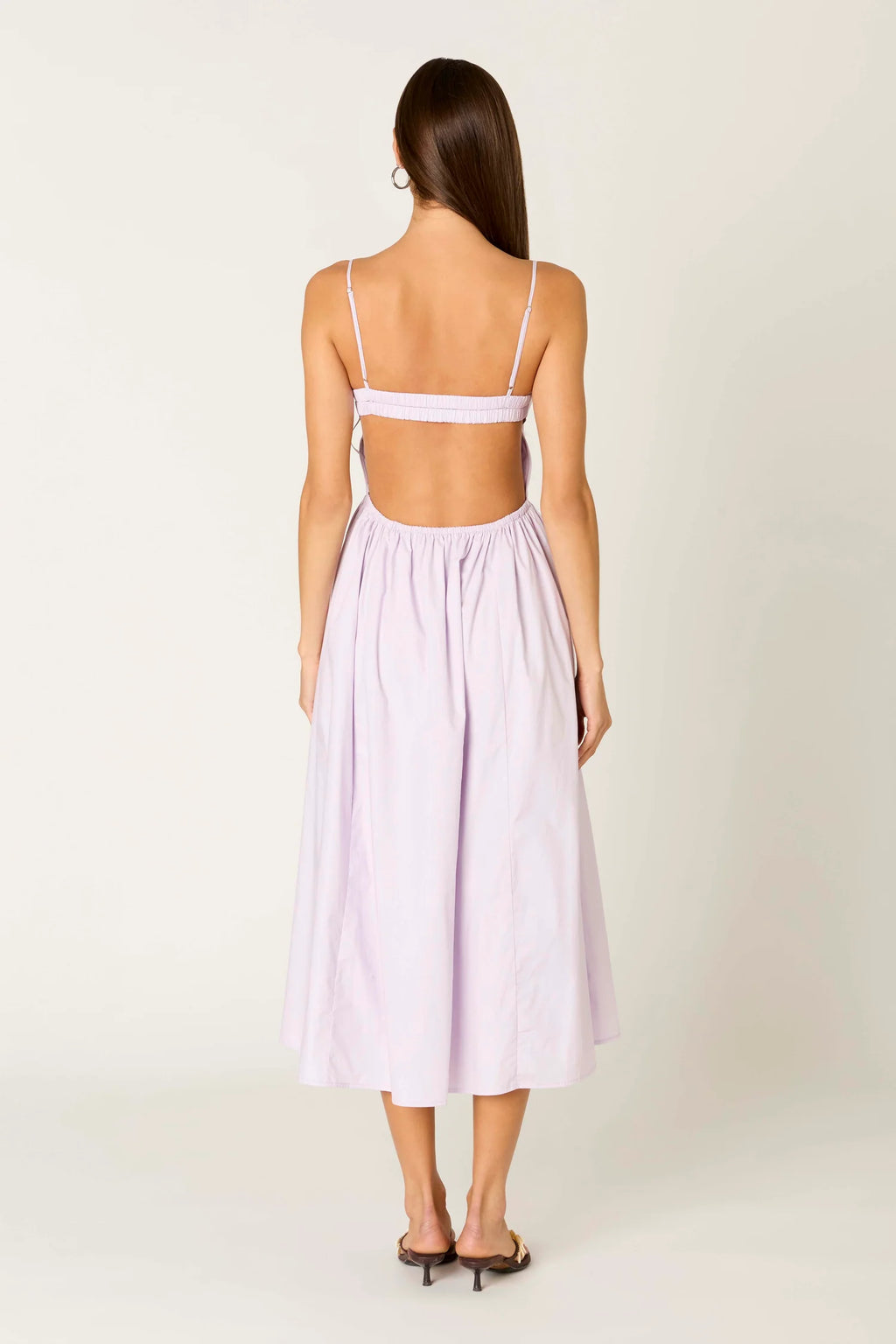 A woman shown from the back wearing a light lavender midi dress with thin shoulder straps, a wide strap across the upper back, and a gathered skirt, photographed against a plain off-white studio background.