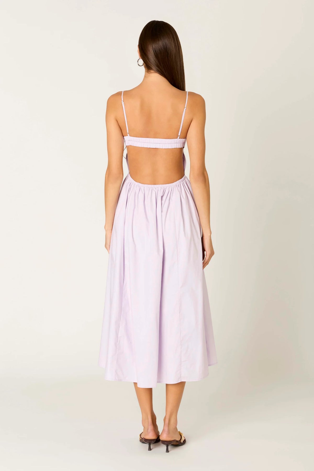 A woman shown from the back wearing a light lavender midi dress with thin shoulder straps, a wide strap across the upper back, and a gathered skirt, photographed against a plain off-white studio background.
