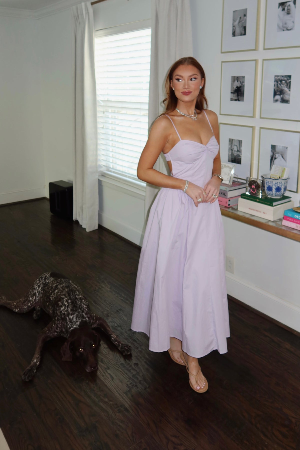 A woman wearing a light lavender spaghetti-strap midi dress stands in a home interior with dark wood floors, white walls, and a dog lying on the floor nearby.