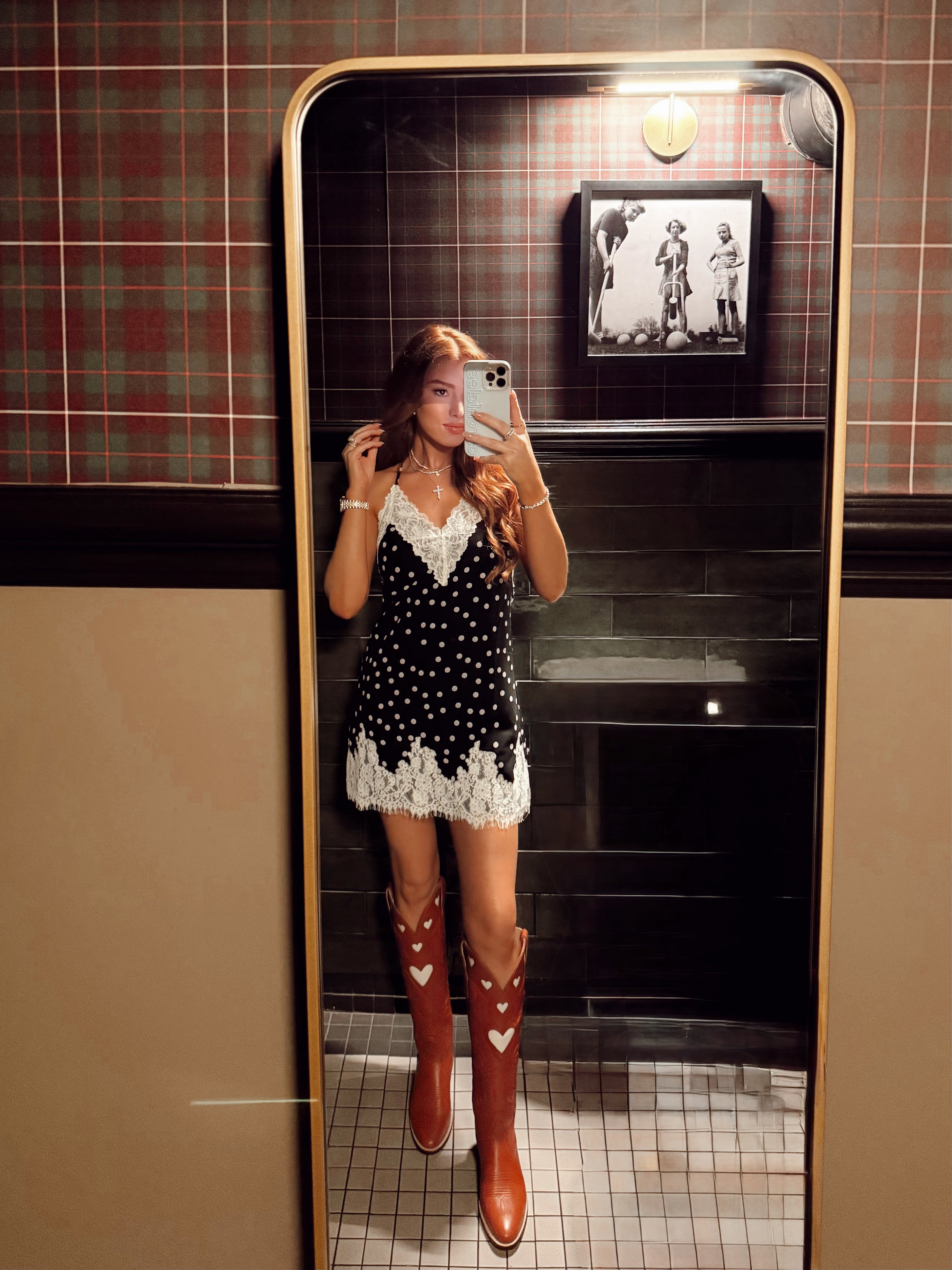 A woman takes a mirror selfie in a bathroom wearing a black and white polka dot mini dress with white lace trim and brown cowboy boots with white heart details.