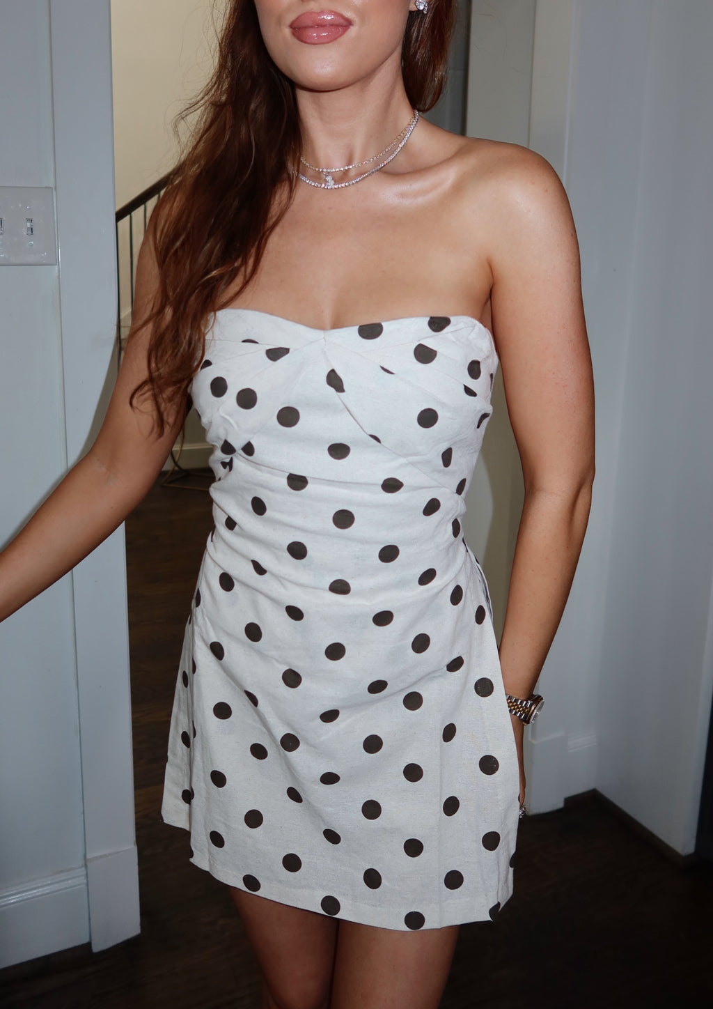 A woman wearing a strapless white mini dress with large dark brown polka dots, standing indoors against a light-colored wall.