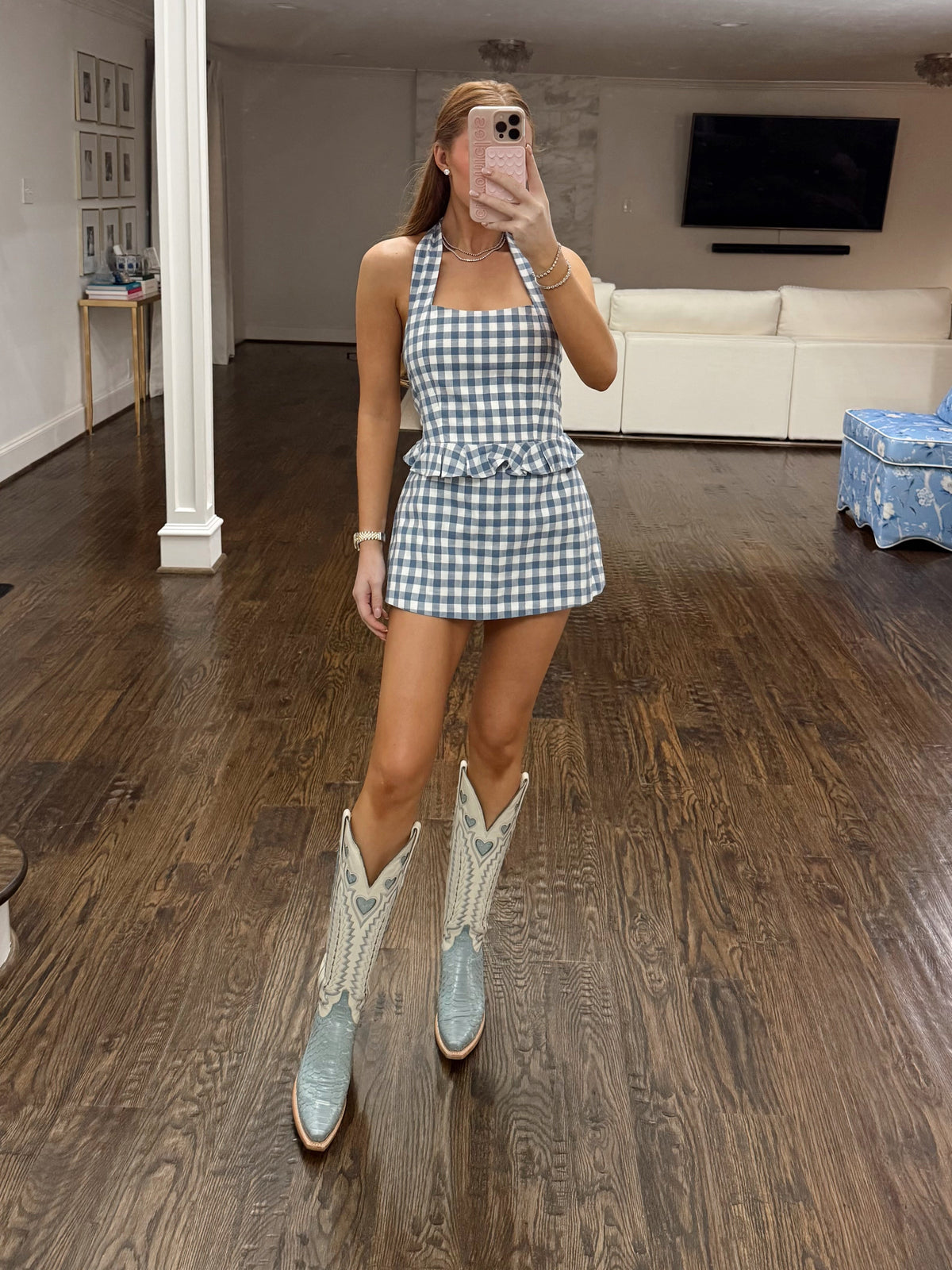 A woman takes a mirror selfie in a spacious home interior, wearing a blue and white gingham halter mini dress paired with light blue western-style cowboy boots with white embroidery.