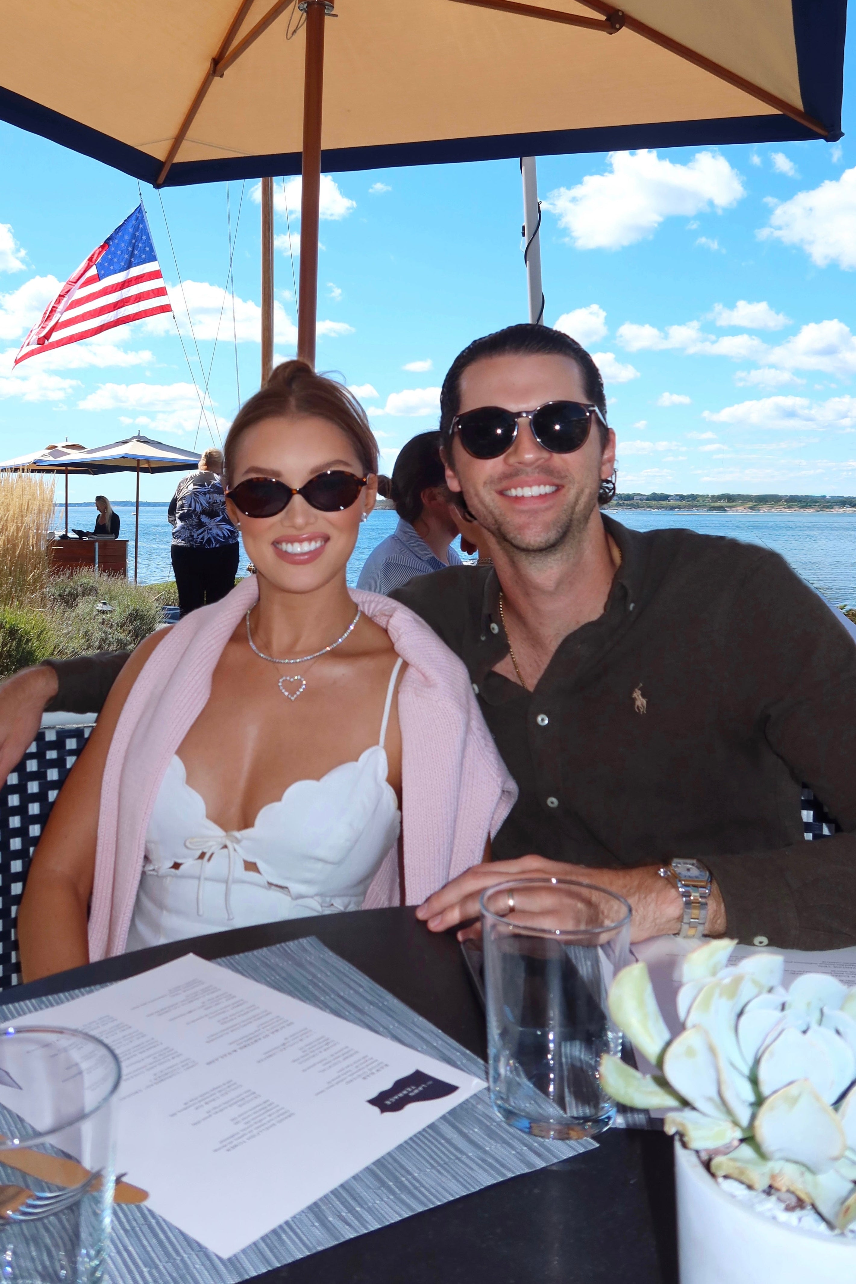 A man and a woman sitting together at an outdoor beachside restaurant table under a large beige patio umbrella. The woman wears a white top and a light pink cardigan, and the man wears a dark brown shirt and sunglasses.
