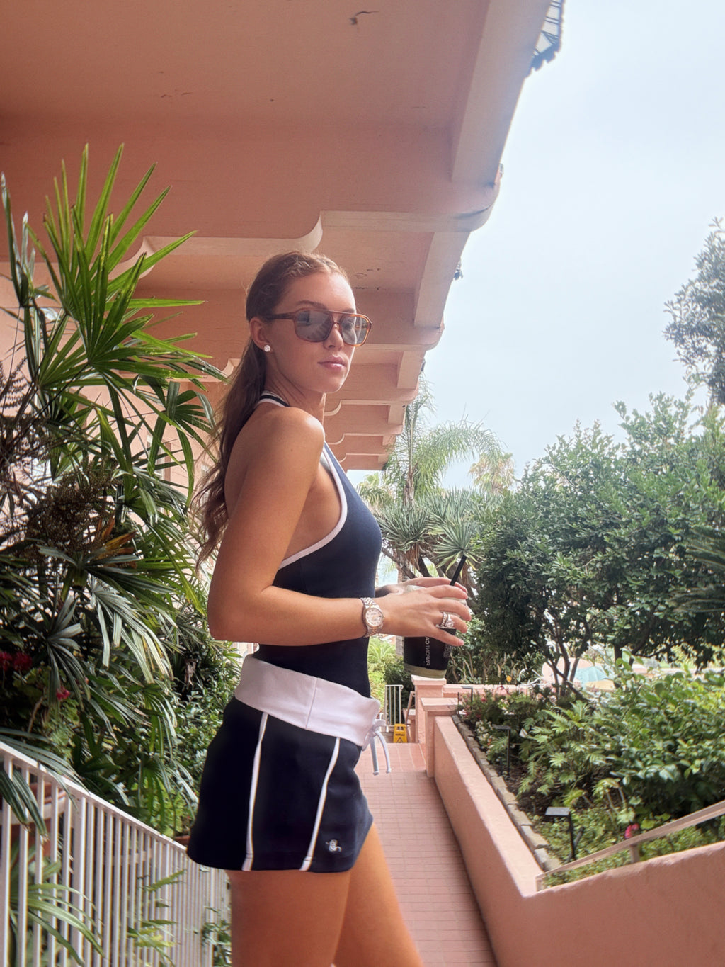 A woman wearing a navy and white athletic tank top and matching shorts stands on an outdoor balcony with tropical plants and a pink structure in the background.