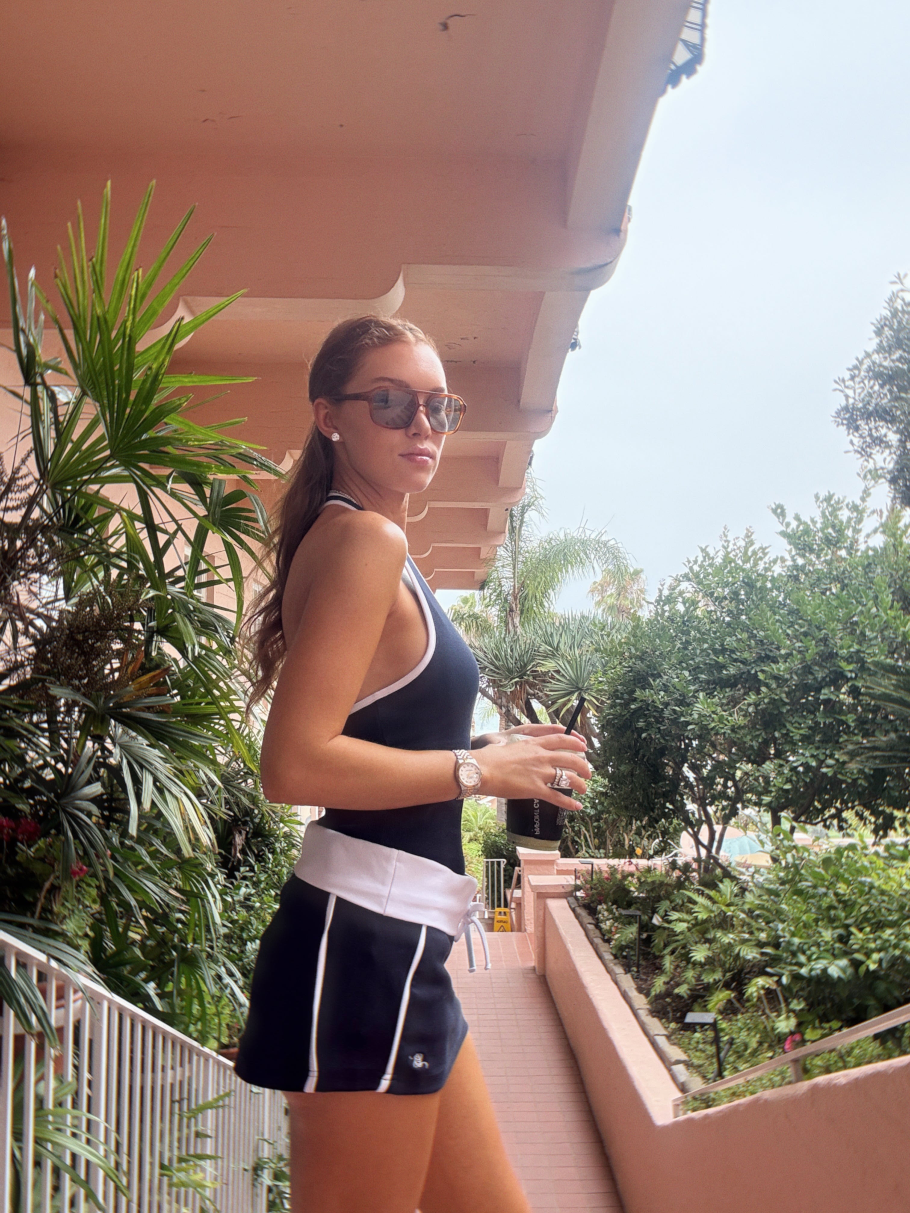 A woman wearing a navy and white athletic tank top and matching shorts stands on an outdoor balcony with tropical plants and a pink structure in the background.