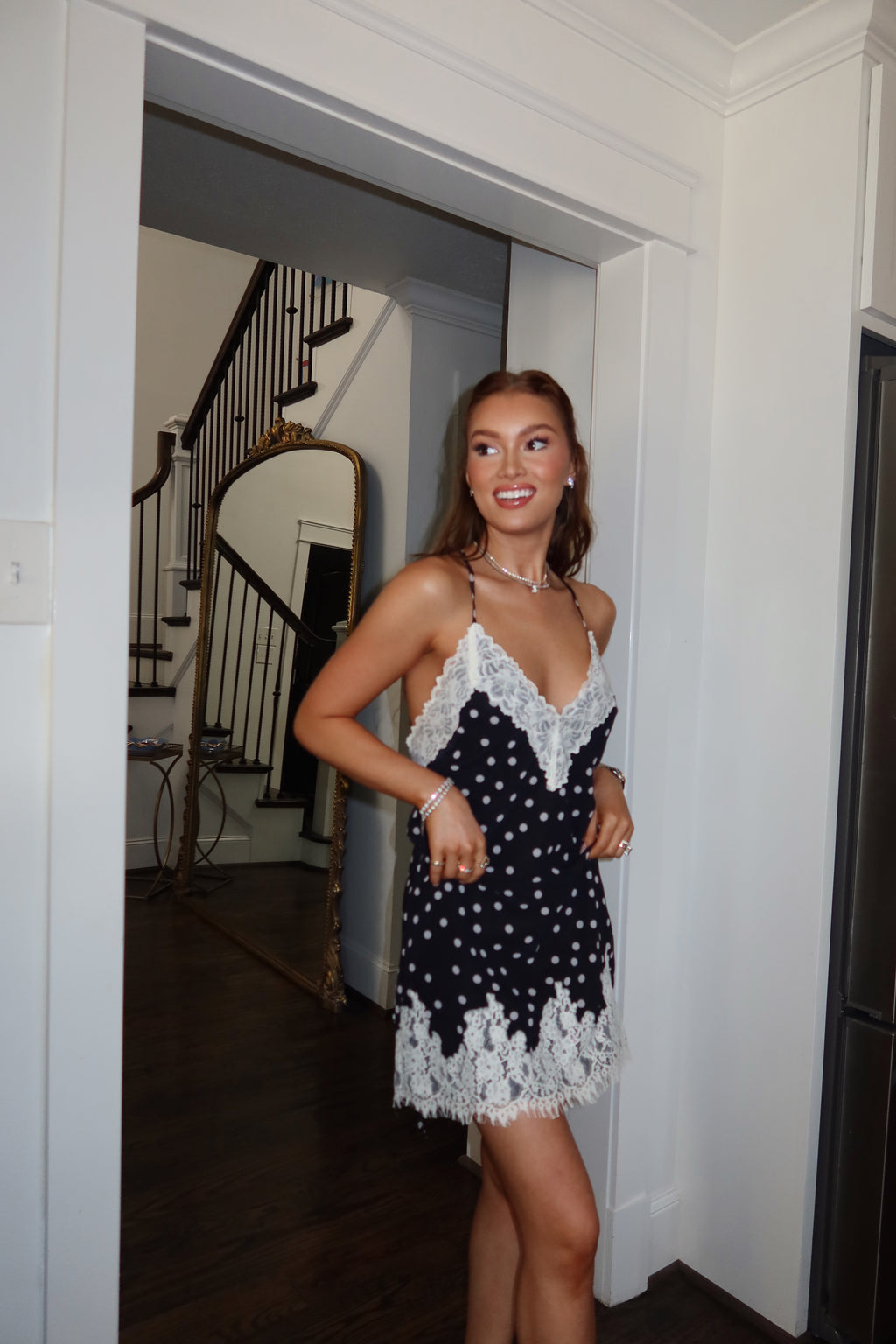 A woman modeling a short black and white polka dot mini dress with white lace trim, standing in a doorway of a home interior.