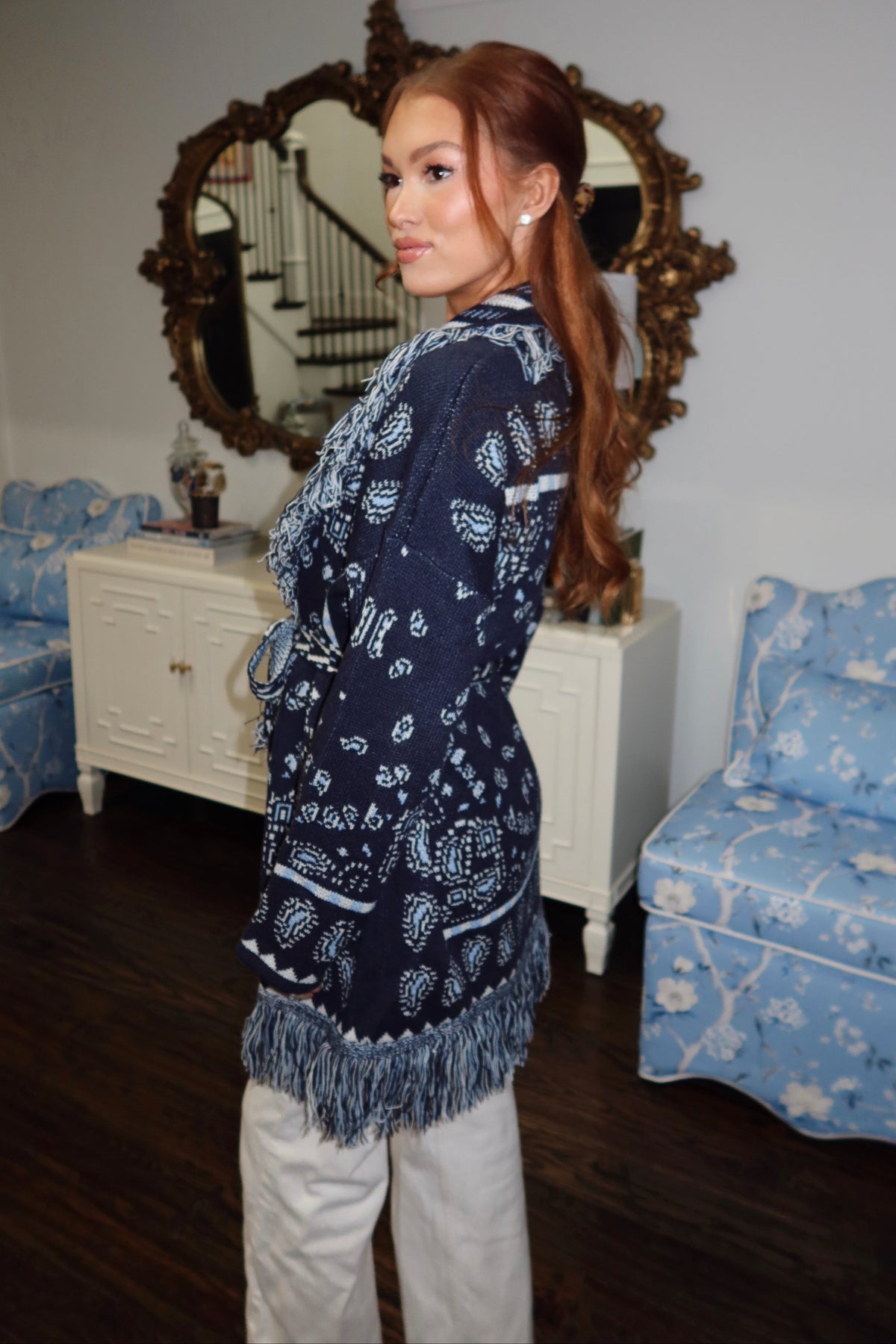 A woman with long red hair is shown from the back in a home interior, wearing a navy blue and white paisley bandana jacket with fringe trim and white pants. The room features a white cabinet, a large ornate gold mirror, and blue patterned armchairs.