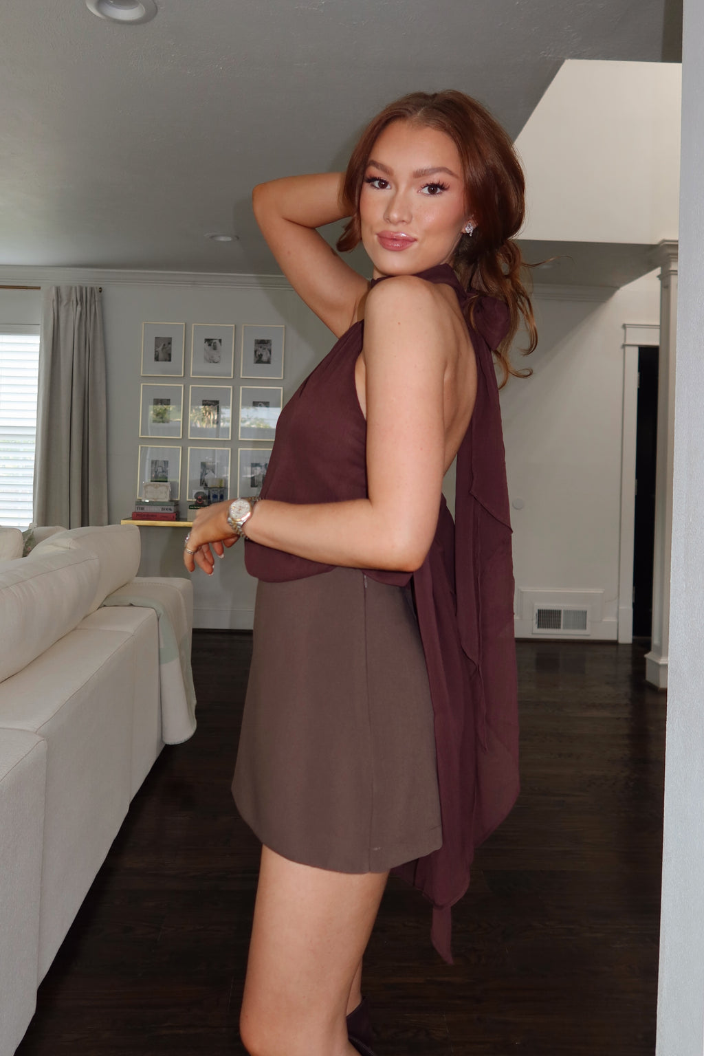 A woman modeling a sleeveless brown mini dress with a draped, ruffled overlay in a home interior setting.