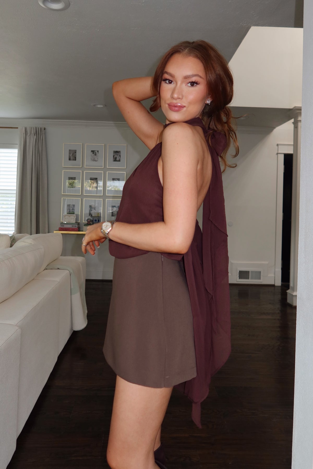 A woman modeling a sleeveless brown mini dress with a draped, ruffled overlay in a home interior setting.