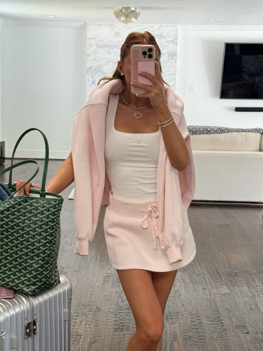 A woman takes a mirror selfie in a modern room, wearing a light pink hooded cardigan over a white ribbed tank top and a matching light pink mini skirt. She is holding a green patterned handbag next to a silver suitcase.