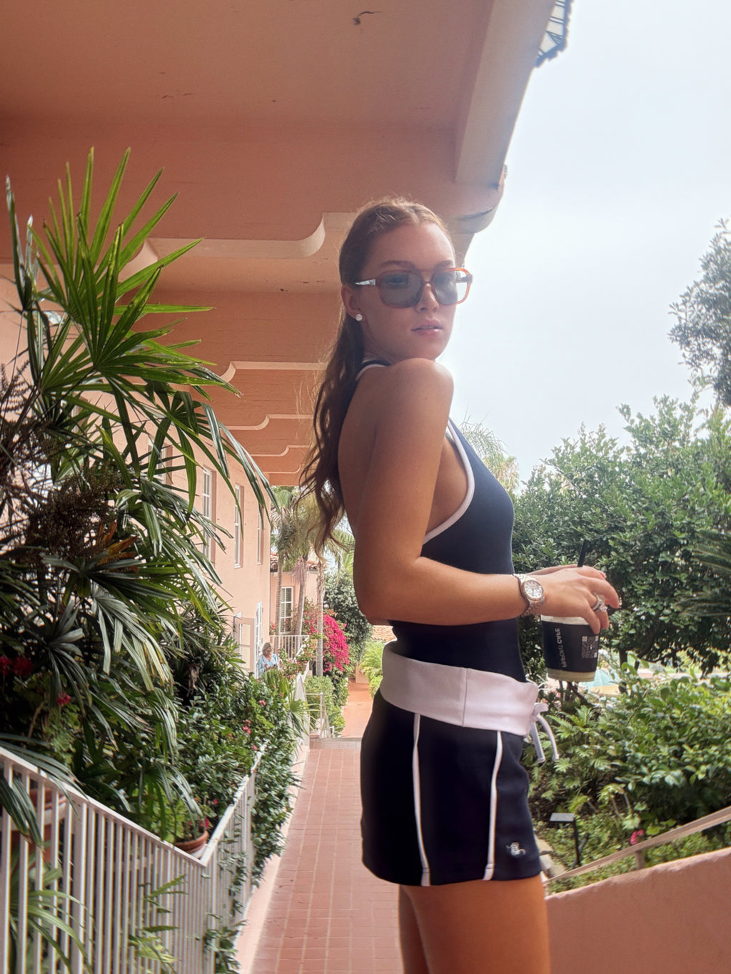 A woman wearing a black athletic tank top and matching shorts with white trim stands on an outdoor balcony holding a black coffee cup. The setting features a terracotta walkway, lush green plants, and a peach-colored building in the background.