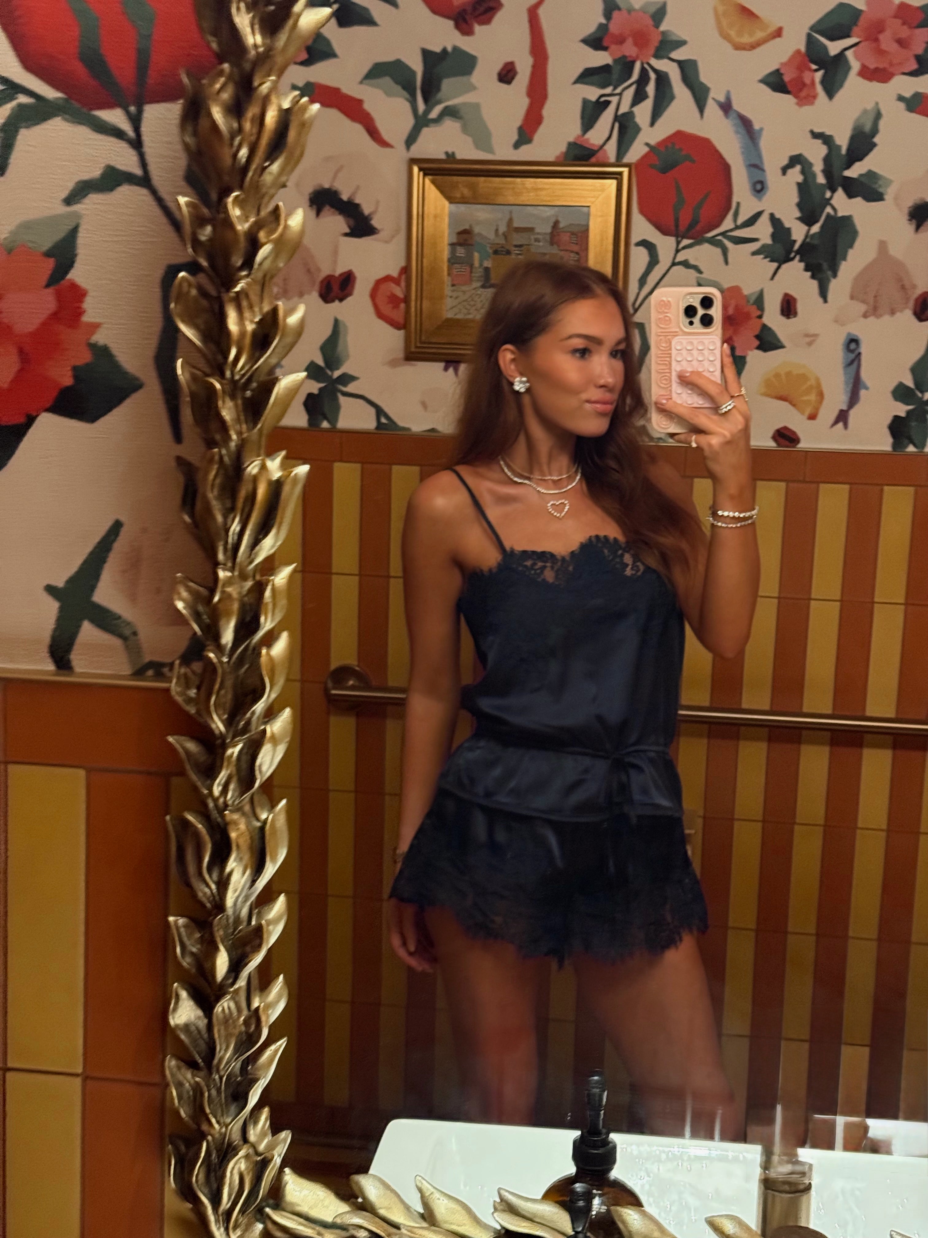 A woman takes a mirror selfie in a bathroom wearing a dark navy blue satin camisole dress with lace trim and a ruffled mini skirt. The background features floral wallpaper, wood paneling, and a large gold mirror frame.